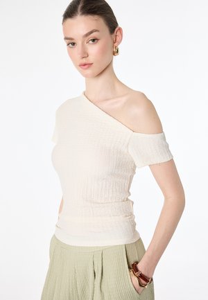 Frau trägt ein cremefarbenes, strukturiertes Off-Shoulder-Top, einen hellgrünen plissierten Rock, goldene Creolen und ein massives braunes Armband, Hand in der Tasche.