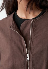 Brown zipped jacket featuring a smooth fabric with a round neckline and a silver zipper, shown against a dark shirt underneath.