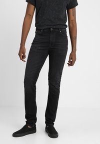 Vaqueros de mezclilla negra con un corte ajustado, diseño de cinco bolsillos y detalles de costura sutiles. Combinados con una camiseta negra y zapatos de slip-on.