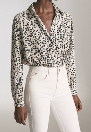 Kvinde iført en beige bluse med lange ærmer og sorte abstrakte pletter, stoppet ned i højtaljede hvide bukser, stående mod en ensfarvet baggrund.