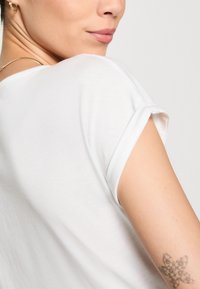 Bílý tričko s ohrnutými rukávy, vyrobené z měkké bavlny. Detailní záběr na límec a rameno, ukazující hladkou strukturu a minimalistický design.