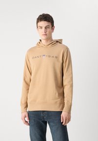Felpa beige con logo frontale, cappuccio con cordini e maniche lunghe. Realizzata in un tessuto morbido con polsini e orlo a coste.