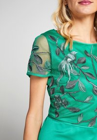 Zelené vyšívané šaty s průhledným topem krátkého rukávu zdobeným listovými a květinovými vzory. Tkanina má hladkou texturu a živou barvu.