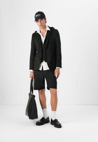 Schwarze Blazer mit Kapuze, weißes Hemd, schwarze Shorts, schwarze Tasche, schwarze Schuhe und weiße Socken. Lässiges modernes Design mit kontrastierenden Texturen.