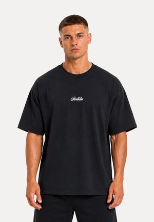 Jeune homme aux cheveux courts portant un t-shirt noir oversized avec un petit texte blanc sur la poitrine, debout devant un fond clair uni.