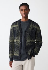 Temno pulover do kolen s zelenim in sivim karo vzorcem, zapenjanjem na gumbe, rebrastimi manšetami in mehko teksturo, nošen preko sive srajce.