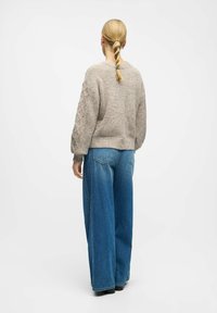 Suéter de punto beige con textura, de corte holgado y dobladillo acanalado, combinado con jeans de mezclilla azul de pierna ancha con bolsillos traseros y un aspecto deslavado.