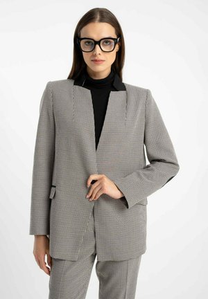 Kobieta w czarnych okularkach i czarnym golfie, ubrana w szaro-białą marynarkę w kratę typu houndstooth oraz dopasowane spodnie.