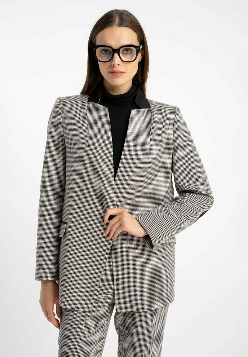 Kobieta w czarnych okularkach i czarnym golfie, ubrana w szaro-białą marynarkę w kratę typu houndstooth oraz dopasowane spodnie.
