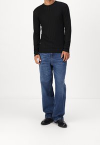 Uomo che indossa una maglietta a maniche lunghe nera a coste, jeans blu larghi e scarpe di pelle nere, in piedi contro uno sfondo bianco semplice.