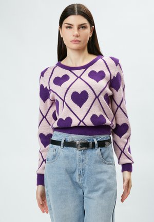 Pull léger rose avec des motifs de cœurs violets et de formes de diamants, complété par un col et des poignets côtelés violets. Ceinture noire avec boucle métallique.