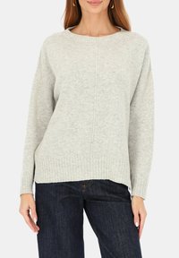 Kobieta ubrana w jasnoszary sweter z dzianiny z prążkowanymi mankietami i krawędzią, w zestawie z ciemnoniebieskimi dżinsami na jednolitym tle.