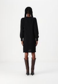 Robe noire en maille avec manches longues bouffantes, longueur genou, avec une fente au dos. Associée à des bottes en cuir marron de style western.
