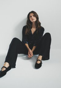 Schwarzes Samt-Pyjama-Set mit langen Ärmeln und weitem Hosenbein, kombiniert mit schwarzen Ballerinas mit einem Riemen und Schnallendetails.