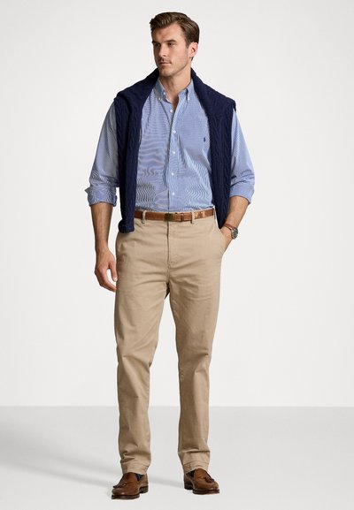 Un gilet en maille bleu marine sur une chemise à rayures bleu clair et blanche à manches longues, un pantalon chino beige, et des mocassins marron avec des détails.
