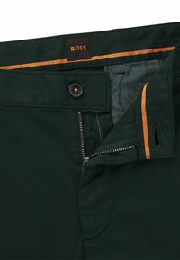 Ciemnozielone spodnie z zamkiem błyskawicznym ozdobionym pomarańczowymi przeszyciami, czarnym guzikiem do zapięcia oraz brązową metką z napisem „BOSS”.