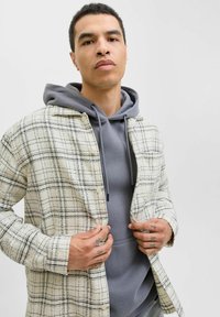 Kariertes Hemd in Creme- und Schwarztönen, über einen grauen Hoodie geschichtet. Verfügt über eine Knopfleiste, zwei Brusttaschen und einen entspannten Schnitt.