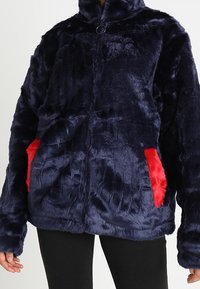 Mörkblå faux fur-jacka med hög krage, försedd med röda accentfickor och dragkedja. Mjuk textur och oversize-passform.