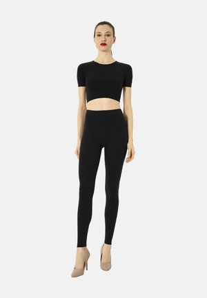 Frau steht, trägt ein schwarzes kurzärmeliges Crop-Top, schwarze Leggings und beige High Heels vor einem hellgrauen Hintergrund.