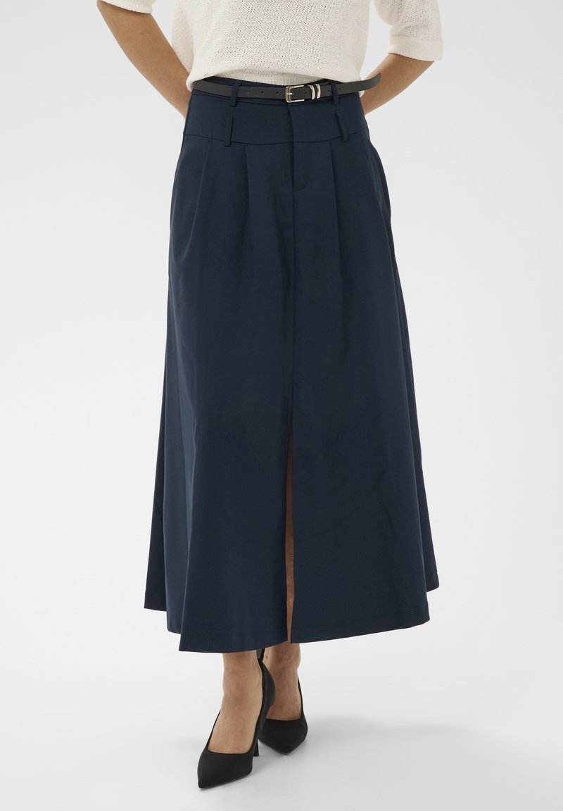 Femme portant une jupe midi bleu marine avec une fente à l'avant et une ceinture noire, associée à des chaussures à talons noirs et un haut blanc.