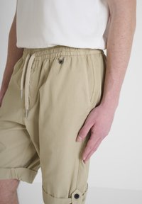 Pantaloni shorts in cotone beige con vita elastica e cordoncino, tasche laterali e polsini con bottone. Tessuto liscio e vestibilità casual.
