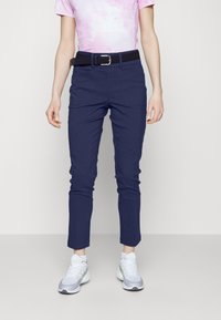 Osoba ubrana w granatowe, dopasowane spodnie, czarny pasek, różową koszulę w stylu tie-dye oraz białe sneakersy, stojąca na tle jasnego, jednolitego tła.