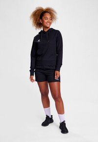 Schwarzer Kapuzenpullover mit Fronttasche, kombiniert mit schwarzen Shorts. Das Modell trägt weiße Socken und schwarze Turnschuhe, die einen sportlichen Look präsentieren.