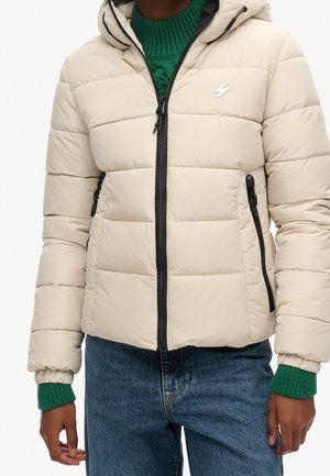 Person wearing beige quilted puffer jacket with black zipper and pockets, green knit sweater cuffs visible, and blue denim jeans.