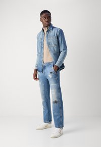 Chaqueta de mezclilla con bolsillos frontales y textura desgastada, combinada con un suéter beige y jeans azules desgastados. Zapatillas blancas completan el conjunto.