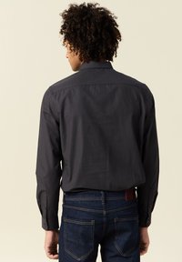 Camicia button-up grigio scuro con maniche lunghe, caratterizzata da una texture liscia e un colletto classico, abbinata a jeans in denim blu. Vista posteriore.