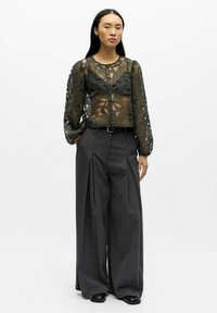 Sheer olive green lace blouse with floral pattern and button closure, paired with high-waisted, wide-leg gray trousers and black shoes.