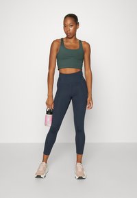 Mörkgrön kort sportbehå, högmidjade marinblå leggings och ljusrosa sneakers. Håller en genomskinlig vattenflaska med rosa detaljer.