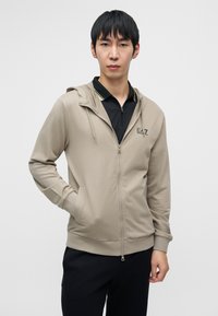 Φωτεινή μπεζ ζακέτα με φερμουάρ και κουκούλα με κορδόνι, με ομαλή υφή και λογότυπο E.A.Z. στο στήθος. Συνδυάζεται με μαύρο πουκάμισο με γιακά.