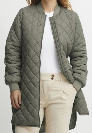 Žena v olivově zelené prošívané bundě rozepnuté, přes bílý top a béžové kalhoty, s rukama částečně v kapsách.