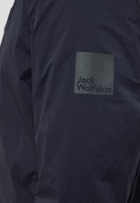 Marineblaue Jacke aus glattem Stoff mit einem quadratischen Logo-Patch in einem helleren Farbton, auf dem "Jack Wolfskin" in fetter Schrift zu sehen ist.