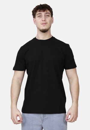 Giovane uomo con capelli corti e ricci che indossa una maglietta nera semplice e jeans azzurri chiari su uno sfondo chiaro uniforme.