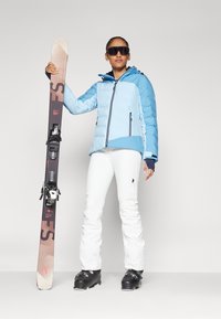 Lichtblauwe geïsoleerde skijal met donkere accenten, witte skibroek en zwarte skischaatsen, houdend bruine skies met bindingen en logo.