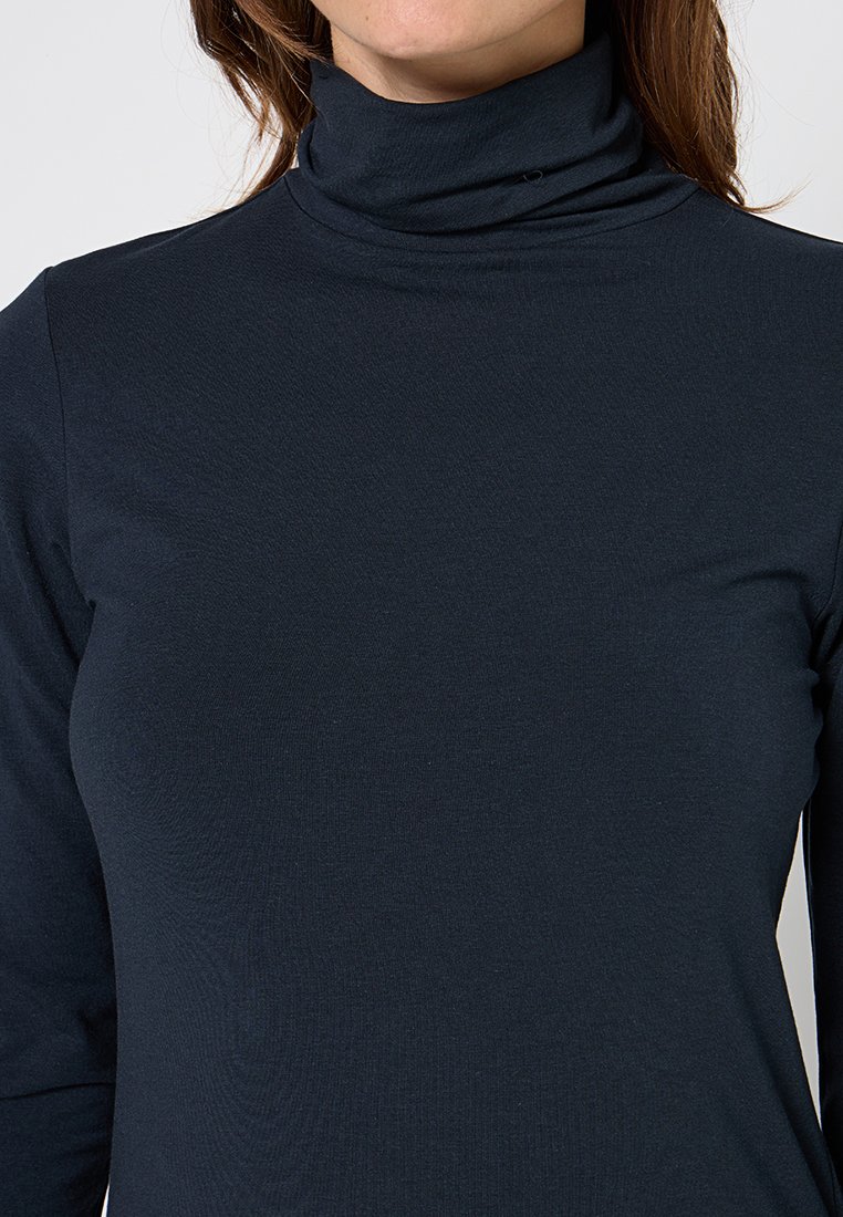 Vrouw in een nauwsluitende zwarte turtleneck trui met lange mouwen, zichtbaar van de schouders tot halverwege de torso voor een effen achtergrond.