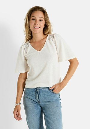 Haut de couleur crème en tissu doux avec un col en V, des manches courtes évasées et des détails en dentelle sur les épaules. Associé à un jean bleu.