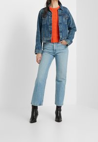 Kobieta ubrana w niebieską dżinsową kurtkę, jasnopomarańczowy sweter w warkocze, jasnoniebieskie dżinsy i czarne botki do kostki, stojąca na białym tle.