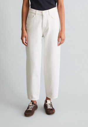 Person, die hoch taillierte weiße Jeans mit geradem Bein und dunkelbraune Turnschuhe mit weißen Schnürsenkeln trägt, steht vor einem hellen Hintergrund.