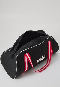 Black sports duffel bag with white piping, red striped handles, and a front zip pocket featuring a white Nike logo.