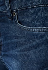 Vaqueros de mezclilla con un tono azul profundo, que cuentan con un bolsillo cosido y un acento de botón plateado. La tela muestra una textura ligeramente desgastada.