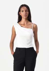 Hvit, ensidig bluse med drapert frontdetalj, laget av glatt stoff. Sammen med svarte skreddersydde bukser, med hendene i lommene.