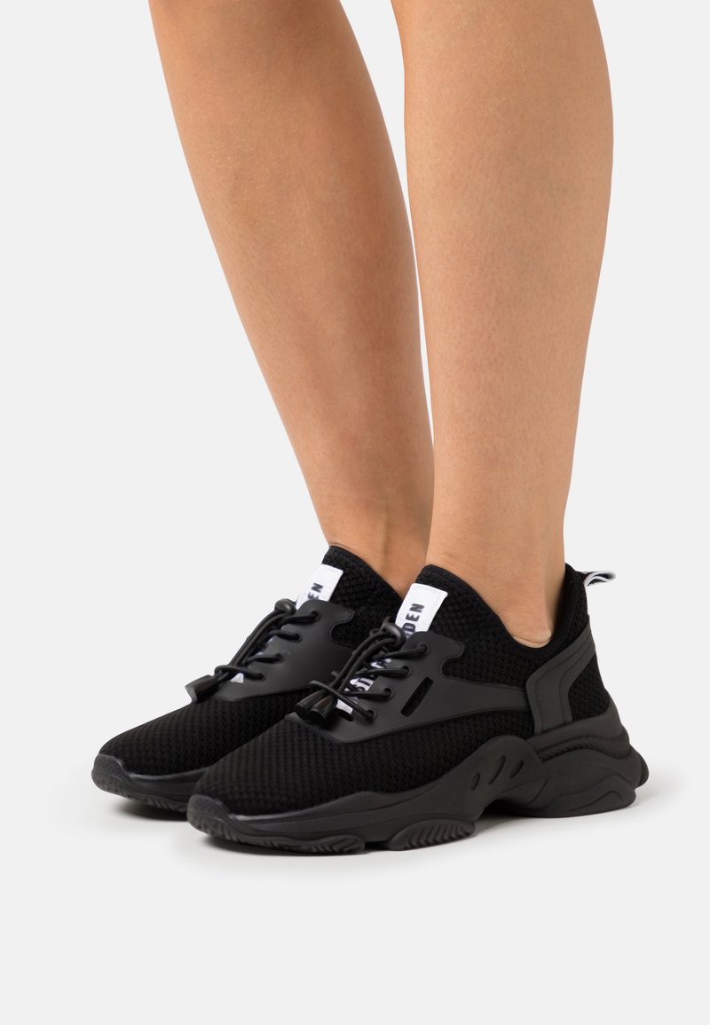 Jambes portant des baskets de sport noires en mesh avec des semelles épaisses et des languettes blanches, debout sur une surface blanche unie.