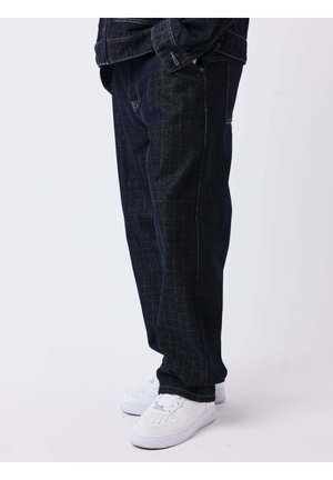 Personne portant un pantalon à carreaux foncés et des baskets blanches, debout les mains dans les poches devant un fond uni.
