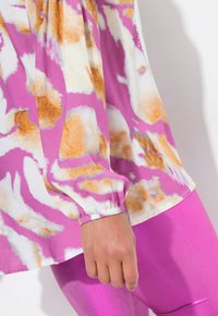 Blusa a maniche lunghe con motivo astratto rosa e bianco con accenti marroni; abbinata a leggings rosa lucidi; polsini elasticizzati visibili.