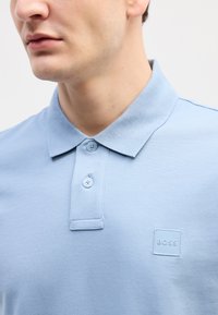 Gaiši zils polo krekls ar teksturētu audumu, divām pogām un kvadrātveida, izcilu logo, kas izšūts uz krūtīm. Tuvplāns.