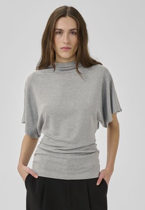 Femeie cu păr lung și castaniu, purtând un tricou cu mâneci scurte gri deschis și pantaloni negri, stând cu mâinile în buzunare, în fața unui fundal simplu.