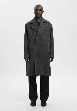 Noor mees, kes kannab üle suure soojalt tumehallist villast mantlit, musta särki, laiu musti pükse ja musti nahast kingi, seisab tavalise tausta ees.